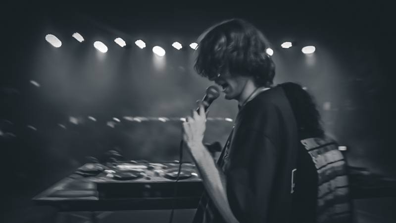 Vocalist performing on stage under stage lights, filmed in black and white